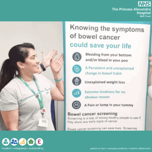 Woman standing beside a newly displayed poster promoting knowing the signs of bowl cancer, smiling and gesturing towards the information.