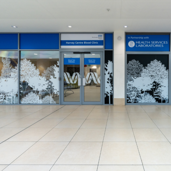Entrance to the new blood clinic with glass double doors labelled “Blood Test Entrance,” blue signage reading “Harvey Centre Blood Clinic,” and frosted window graphics of trees on either side. The space is clean and well-lit, with tiled flooring in front.